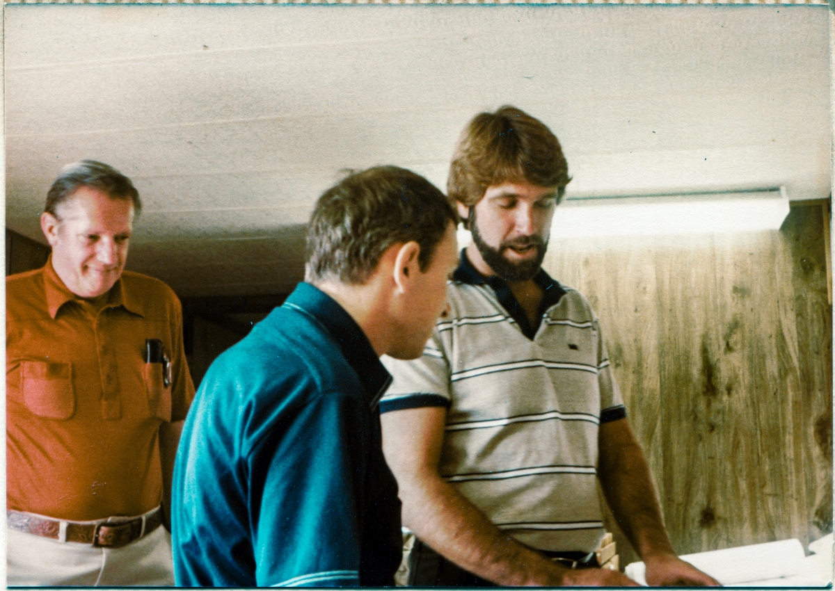 Image 011A. Dick Walls, Gene Hajdaj, and Chris Miller discuss an issue at the drawing table in Sheffield Steel’s field Trailer at Space Shuttle Launch Complex 39-B, Kennedy Space Center, Florida. Dick Walls was Sheffield Steel’s project manager at the Pad. Gene Hajdaj was a structural engineer working for Reynolds Smith & Hills, the A&E firm that did the design and engineering for the Rotating Service Structure which Sheffield fabricated and delivered the structural steel for. Chris Miller was a mechanical engineer, also working for RS&H out at the Pad. Photo by James MacLaren.